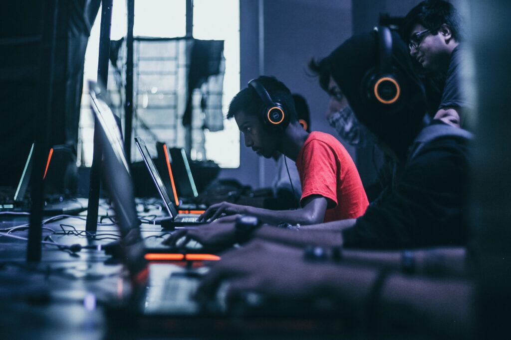 This photo shows a row of middle-school-aged children sitting at laptops with headsets on. The color palate is dark with neon orange lights on the headsets and laptops making it appear very modern.